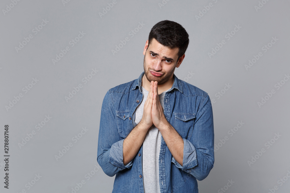 Handsome stylish unshaven young man in denim jeans shirt posing isolated on grey wall background studio portrait. People sincere emotions lifestyle concept. Mock up copy space. hands folded in prayer. Handsome stylish unshaven young man in denim jeans shirt posing isolated on grey wall background studio portrait. People sincere emotions lifestyle concept. Mock up copy space. hands folded in prayer.
