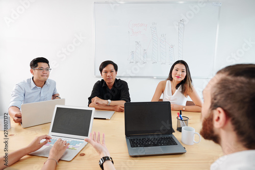 Asian business colleagues sitting at the table and listening to their male leader attentively during business meeting