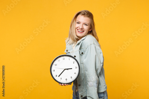 Portrait of smiling young woman in denim casual clothes looking camera, holding round clock isolated on bright yellow orange wall background in studio. People lifestyle concept. Mock up copy space.