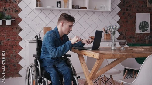 Young and disabled man sitting in special comfort wheelchair and spending his holiday at home with bright light room in modern interior flat. Male using personal computer or laptop and working
