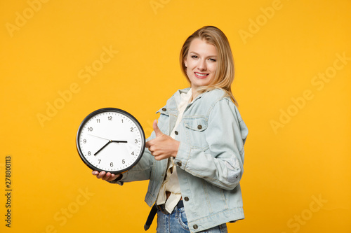 Portrait of smiling young woman in denim casual clothes showing thumb up, looking camera, holding round clock isolated on yellow orange wall background. People lifestyle concept. Mock up copy space.