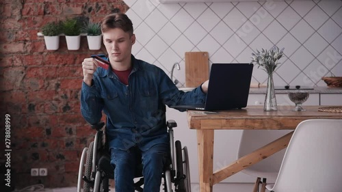 Online shopping concept. Young and disabled man sitting in comfort wheel chair and spending weekend at cozy bright room in house with modern interior. Male using bank card and pc laptop