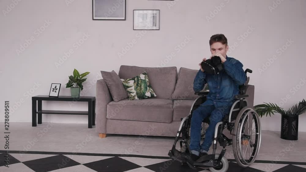 Young and disabled man sitting in special comfort wheelchair and spending free time at cozy house with modern and bright interior room. Male making fun and using virtual reality glasses Young and disabled man sitting in special comfort wheelchair and spending free time at cozy house with modern and bright interior room. Male making fun and using virtual reality glasses