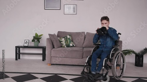 Young and disabled man sitting in special comfort wheelchair and spending free time at cozy house with modern and bright interior room. Male making fun and using virtual reality glasses photo