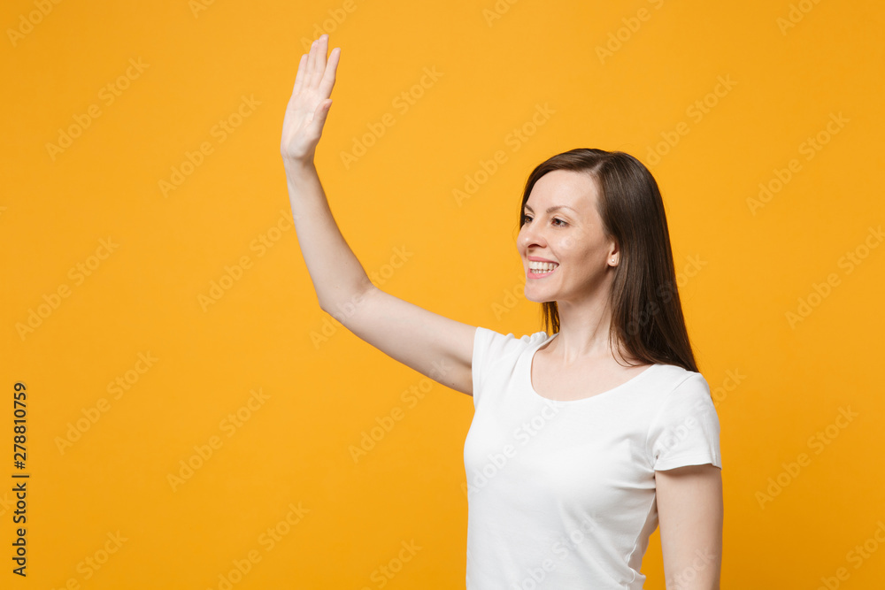 Smiling young woman in white casual clothes looking aside, waving and greeting with hand as notices someone isolated on bright yellow orange background. People lifestyle concept. Mock up copy space. Smiling young woman in white casual clothes looking aside, waving and greeting with hand as notices someone isolated on bright yellow orange background. People lifestyle concept. Mock up copy space.