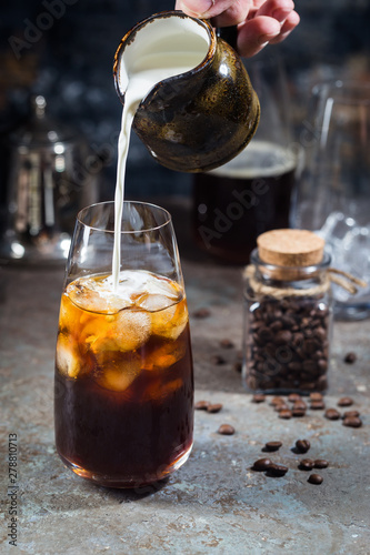 Ice coffee cold summer drink in a tall glass with pouring cream and coffee beans on a stone background.
