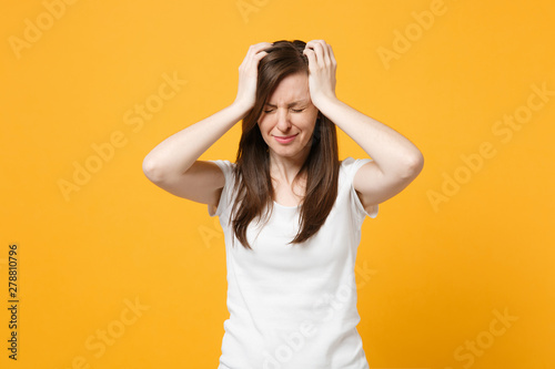 Portrait of displeased crying young woman in white casual clothes keeping eyes closed, putting hands on head isolated on bright yellow orange background. People lifestyle concept. Mock up copy space.