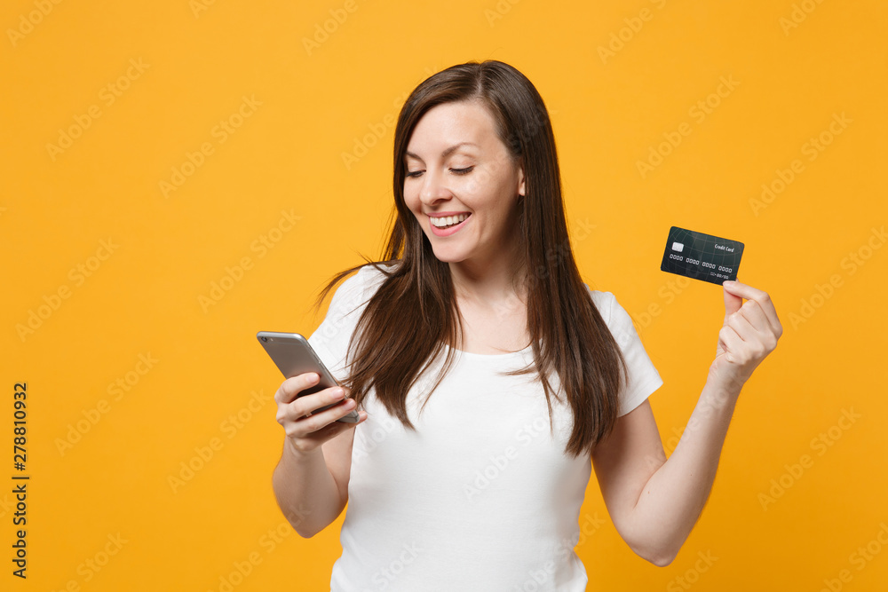 Portrait of smiling young woman in white casual clothes using mobile phone, holding credit bank card isolated on yellow orange wall background in studio. People lifestyle concept. Mock up copy space. - obrazy, fototapety, plakaty 