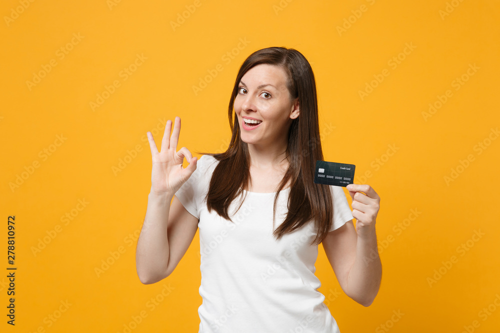Portrait of pretty young woman in white casual clothes showing OK gesture, holding credit bank card isolated on yellow orange wall background in studio. People lifestyle concept. Mock up copy space. - obrazy, fototapety, plakaty 