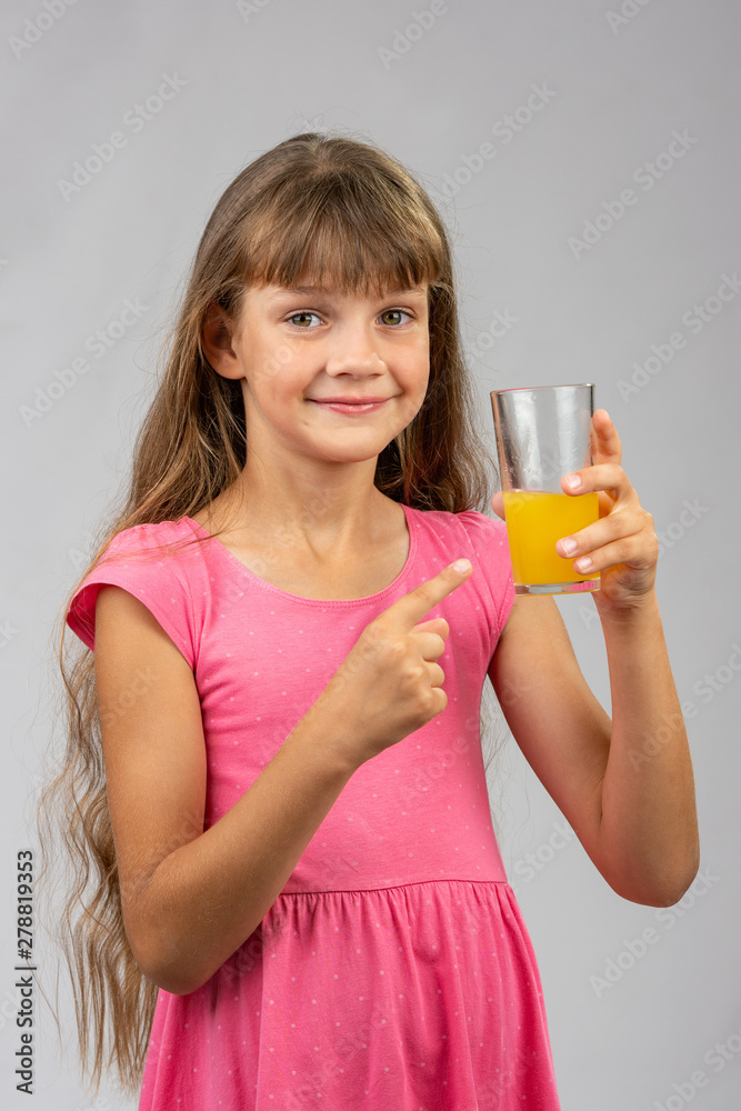 A girl holds a glass of orange juice in her hand and points her finger at it A girl holds a glass of orange juice in her hand and points her finger at it