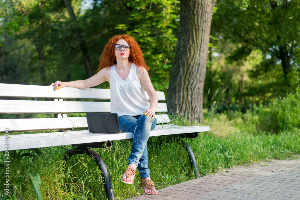 Naklejka premium Red-haired female freelancer working on a laptop while sitting on a park bench.
