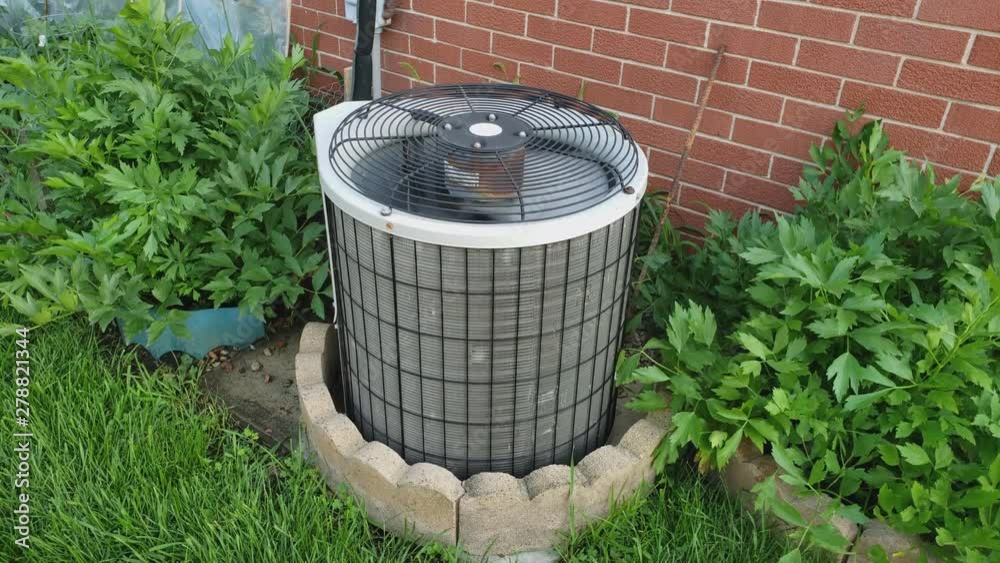Single family home old rusty AC unit or air conditioner external heat exchange unit spinning and blowing air against some plants and brick wall.