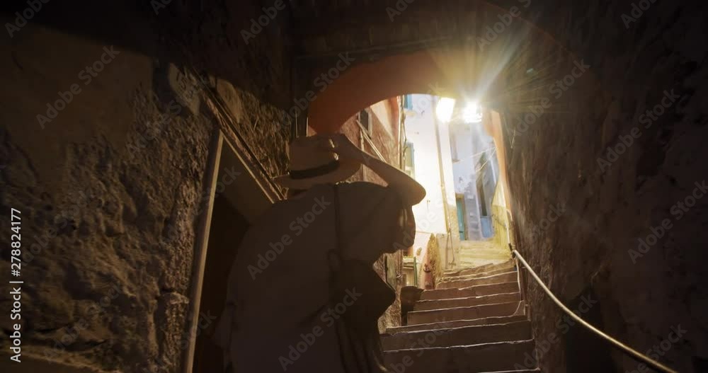 Back view cheerful girl in trendy outfit walking up stone stairs in the beautiful narrow street to the sunlight. Exploring Tuscany, Riomaggiore in summer.