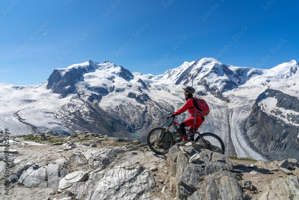 active senior woman, riding her electric mountainbikeon the Gornergrat in Zermatt, Wallis,Switzerland. In The background Gorner Glacier, Monte Rosa, Liskam am Breithorn