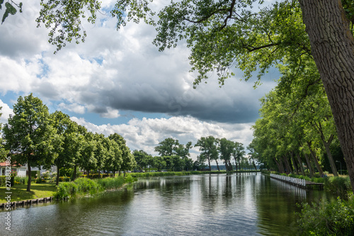 Sluis, the Netherlands - June 16, 2019: Reflecting water of canal to Damme in Belgium, with curtains of green trees on sides, under a storm approaching cloudscape.