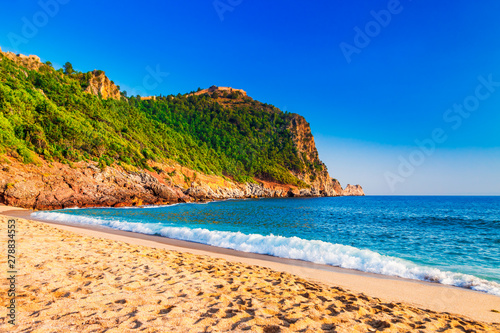 Cleopatra beach on sea coast with green rocks in Alanya peninsula  Antalya district  Turkey. Beautiful sunny landscape for tourism with clear water and sand. Alanya Castle on the cliff