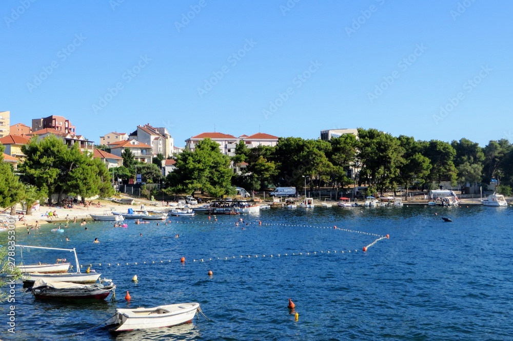 A beautiful amazing view of a beach on Otok Ciovo or Ciovo island beside Trogir, Croatia.  It is a beautiful sunny day with many people swimming in the water and small boats anchored in the bay.