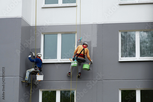 Worker hanging on rope and paints building wall with roller. Painter hanging on cable with paint buckets, industrial climber repairing house facade. Industrial alpinist and climbing. Rigging equipment