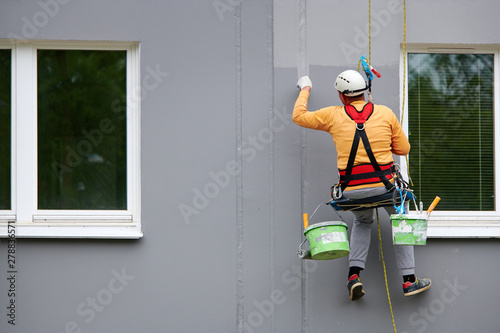 Worker hanging on rope and paints building wall with roller. Painter hanging on cable with paint buckets, industrial climber repairing house facade. Industrial alpinist and climbing. Rigging equipment