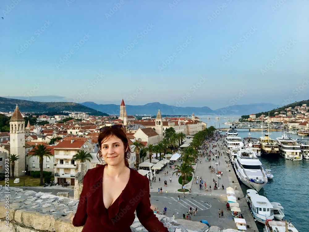 A young female tourist at the top of the Tower Kamerlengo Trogir in the old town of Trogir, Croatia, with the beautiful town and the adriatic sea in the background on a beautiful evening.