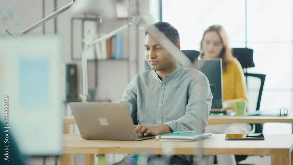 Smart and Handsome Indian Information Technology Specialist Sitting at His Desk works on a Laptop. In the Background Modern Office with Diverse Team of Young Professionals Working Smart and Handsome Indian Information Technology Specialist Sitting at His Desk works on a Laptop. In the Background Modern Office with Diverse Team of Young Professionals Working