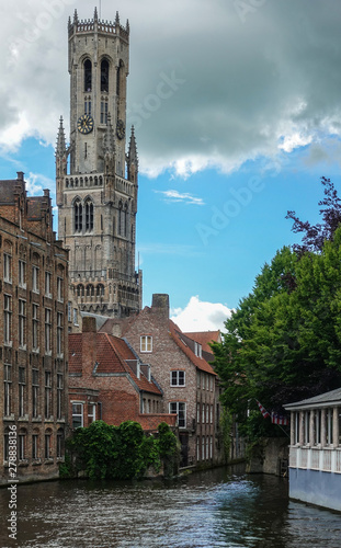 Bruges, Flanders, Belgium -  June 16, 2019: Shot of historic Brugge with Belfry across canal from Huidevettersplein. Brown stone buildings, green foliage and heavy cloudscape. photo