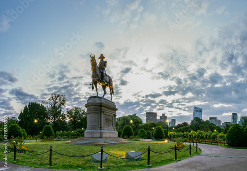 Statue of George Washing stands illuminated under park and dawn lights with downtown Boston in distance.