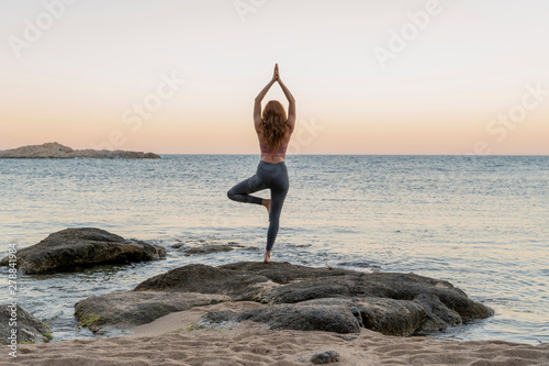 Young woman practicing yoga on the beach, doing tree pose, during sunset in calm beach, Costa Brava, Spain photo