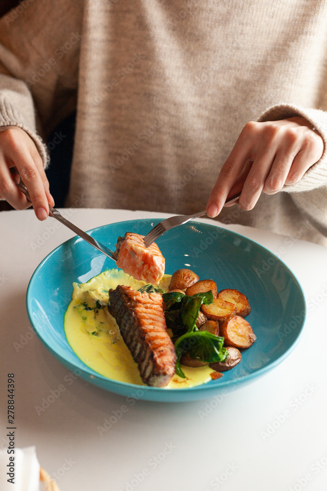 Woman is having a dietary light meal with grilled salmon steak, baked baby potatoes, spinach and coconut milk gravy served on a wooden table