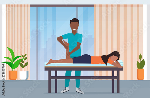 woman lying on massage table african american masseur therapist doing healing treatment massaging patient injured leg physical therapy rehabilitation concept hospital office interior horizontal