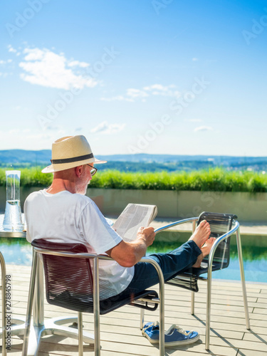 Rear view of senior man reading newspaper, sitting at swimming pool © Westend61