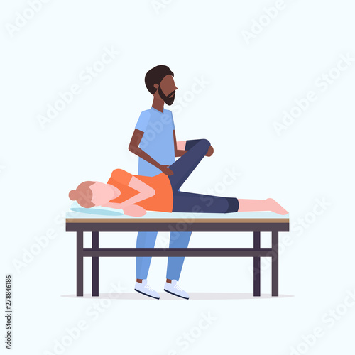 young girl lying on massage bed african american masseur therapist doing healing treatment massaging patient treating legs manual sport physical therapy concept full length