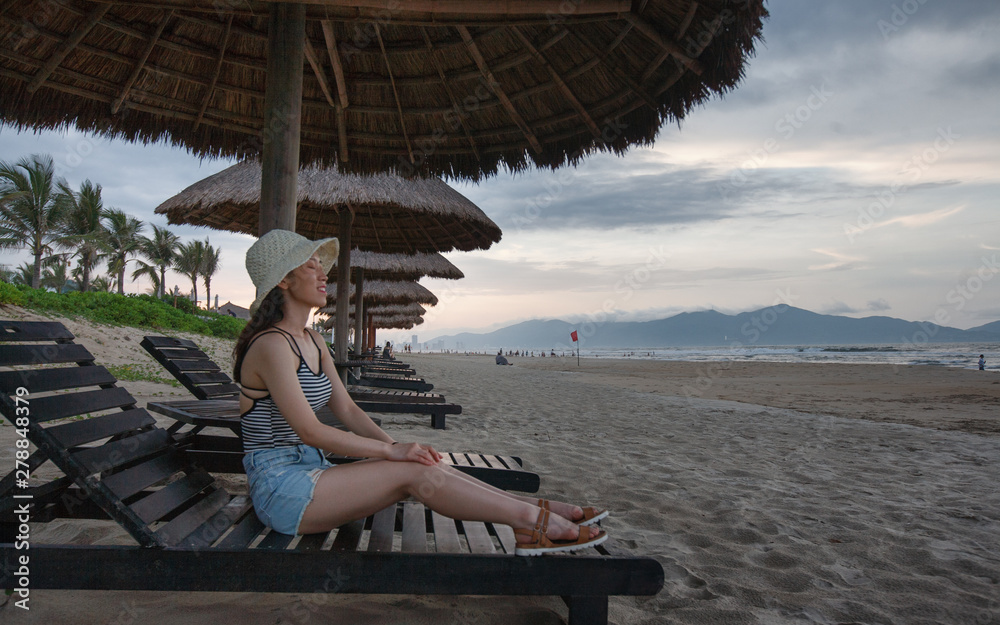 Asian Female Sitting On Beach Recliner with Thatch Umbrella Above