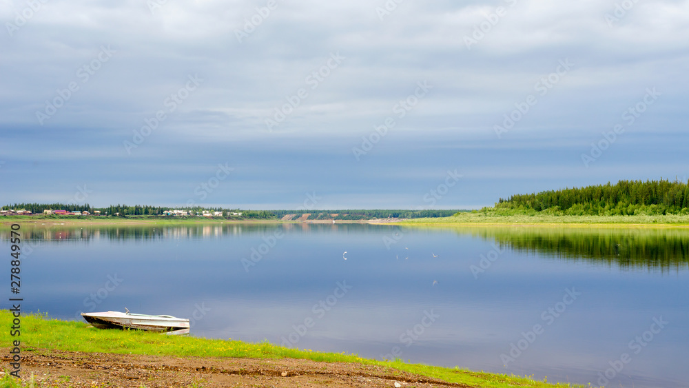 Seagulls fly about a small boat stands on the shore of the Northern Yakut river vilyu near the village of ulus Suntar with houses along the water under a cloudy sky.