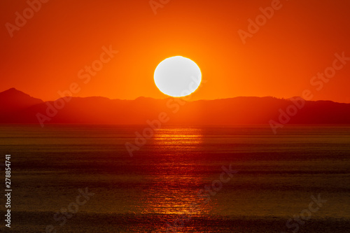 Fototapeta Naklejka Na Ścianę i Meble -  Beautiful golden sunset/sunrise over the sea behind mountains in background. Light reflection on the water as the sun sets/rises. Harmony and beauty in nature. Scenic peaceful ocean view.