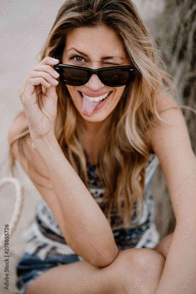 Close-up portrait of pretty blonde woman in black sunglasses sitting on sand and looking at camera sticking out tongue and winking Close-up portrait of pretty blonde woman in black sunglasses sitting on sand and looking at camera sticking out tongue and winking