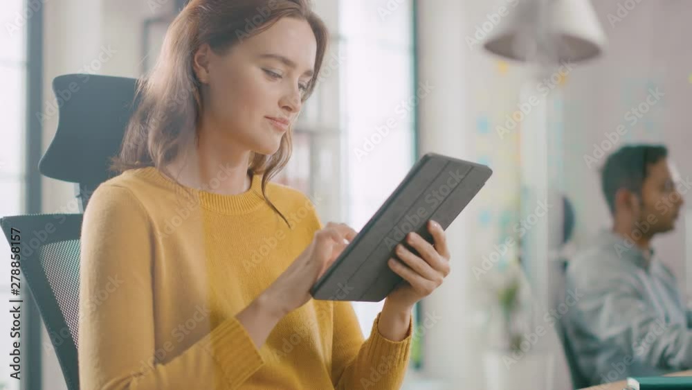 Talented Female Designer Sitting at Her Desk She's Holding and Using Touch Screen Digital Tablet Computer. Bright Office where Diverse Team of Young Professionals Work