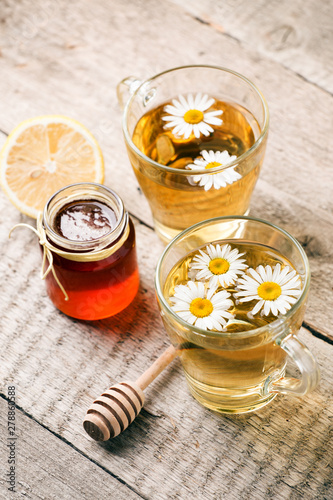Healthy chamomile tea poured into glass cup. Teapot, small honey jar, heather bunch and glass jar of daisy medicinal herbs.