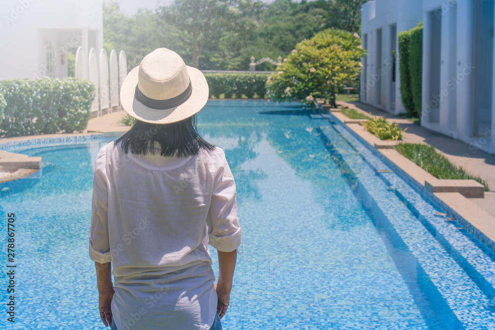 Summer Vacation and Holiday Concept : Woman wear white shirt and weave hat, she standing relax on edge of swimming pool.