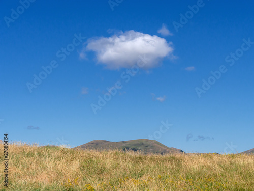 Mountain top grass meadow and bright blue hot summer sk iwth single cloud above. Background.