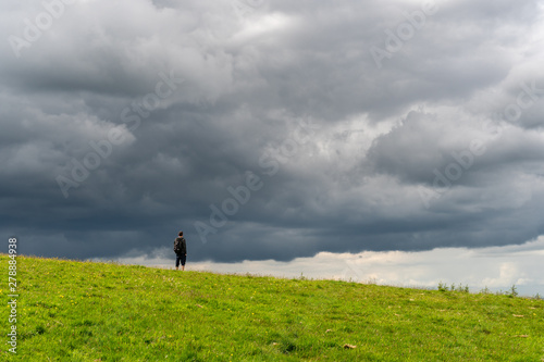 Man hiker standing on a hill top looking at the dark stormy clouds approaching. Chasing summer storms. Outdoor hiking adventure. Feeling small in the Face of the powerful Nature.