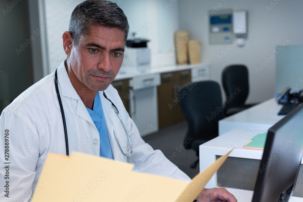 Male doctor looking at medical report while working on computer at desk Male doctor looking at medical report while working on computer at desk