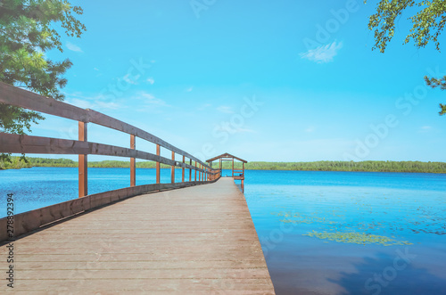 Pier on the lake with calm water under a bright blue sky. Soothing calm minimalistic landscape. Trees surround the pond. Place for text.