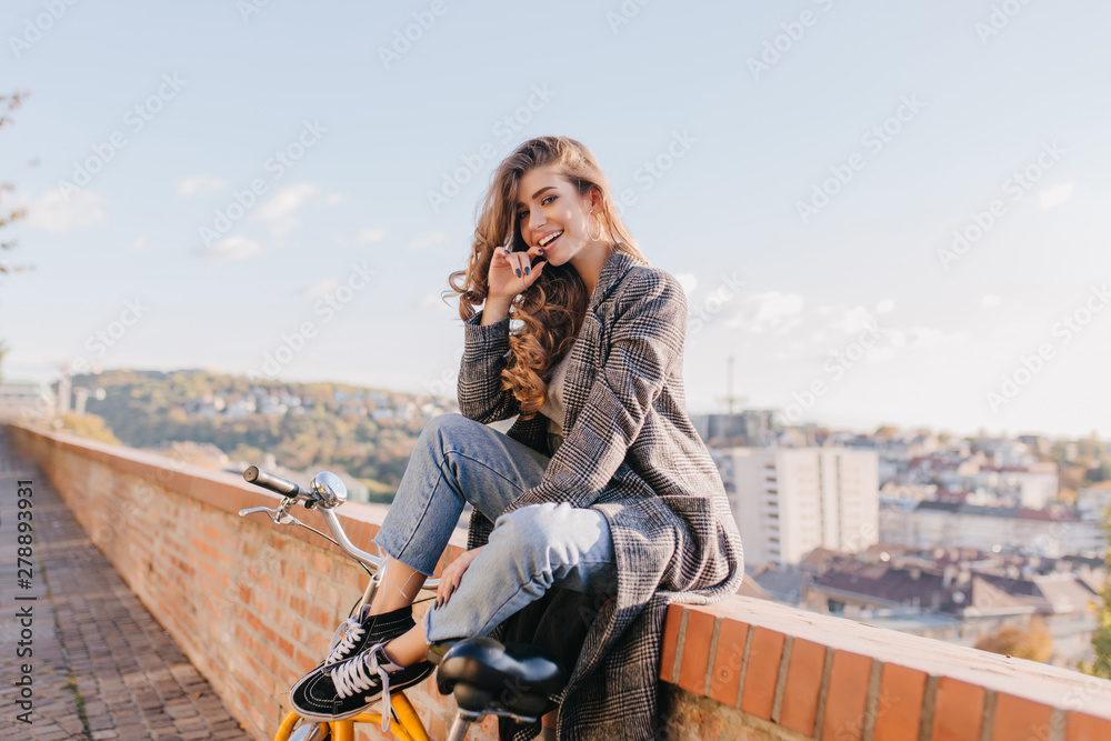 Fototapeta premium Well-dressed lady in good mood playfully posing on city background in warm weather. Charming dark-haired girl in coat sitting on the street under blue sky.