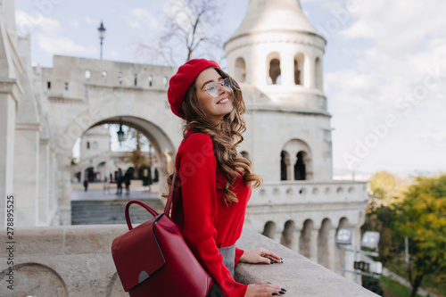Engaging curly woman in red beret enjoying sightseeing during european journey. Outdoor portrait of winsome girl in glasses standing near white palace with smile.