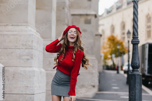 Emotional female model in soft red sweater posing in front of old white building. Portrait of graceful european girl with long curly hairstyle having fun during outdoor photoshoot. © Look!
