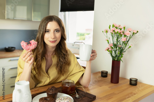 Young beautiful girl is having breakfast at home in the kitchen. She drinks her morning coffee and with great pleasure eats a donut with pink icing.