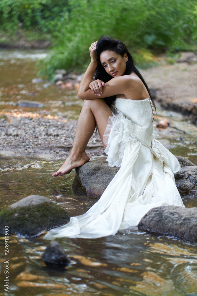 Young pretty brunette woman in white wedding dress sitting on the rock near forest river Young pretty brunette woman in white wedding dress sitting on the rock near forest river