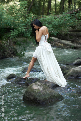 Young pretty brunette woman in white wedding dress trying to cross the river by rocks