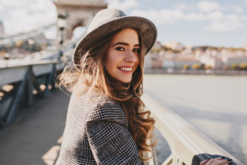 Outdoor photo of romantic european woman with curly hairstyle spending time outdoor, exploring european city. Graceful young lady in gray coat and hat enjoying views on embankment.